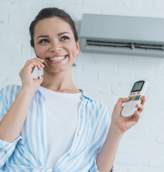 Smiling woman on phone holding ductless mini-split remote, standing near air conditioning unit in bright indoor setting, representing HVAC service satisfaction and customer support.
