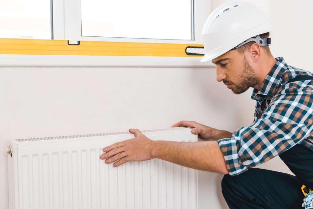 Handyman in a hard hat inspecting a heating radiator, emphasizing heating system maintenance and repair services in a home setting.
