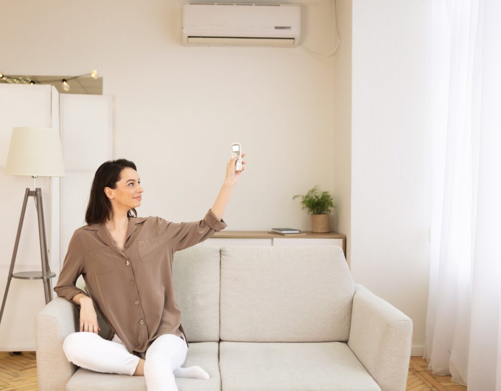 Young woman sitting on a sofa using a remote control to operate an air conditioning unit in a bright living room, illustrating comfort and HVAC service relevance.